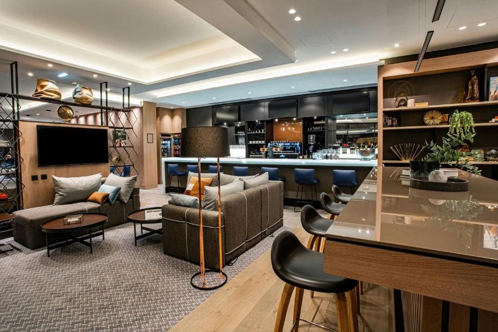 Modern lounge with cozy seating, a large sectional sofa, armchairs, coffee tables, and a high counter with bar stools. Shelves with decor and plants line the walls, and a bar area is visible in the background.