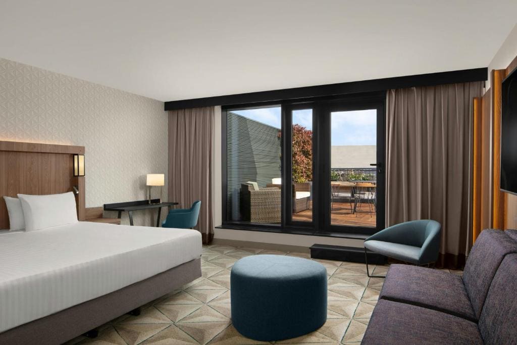 Modern hotel room with a large bed, a desk with a lamp and chair, a lounge chair, ottoman, sofa, and a patio door opening to an outdoor seating area on a terrace. Neutral colors and contemporary decor.