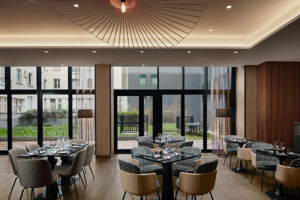 A modern restaurant dining area with large windows, round tables set with glassware, patterned chairs, soft lighting, and a view of a green outdoor space.