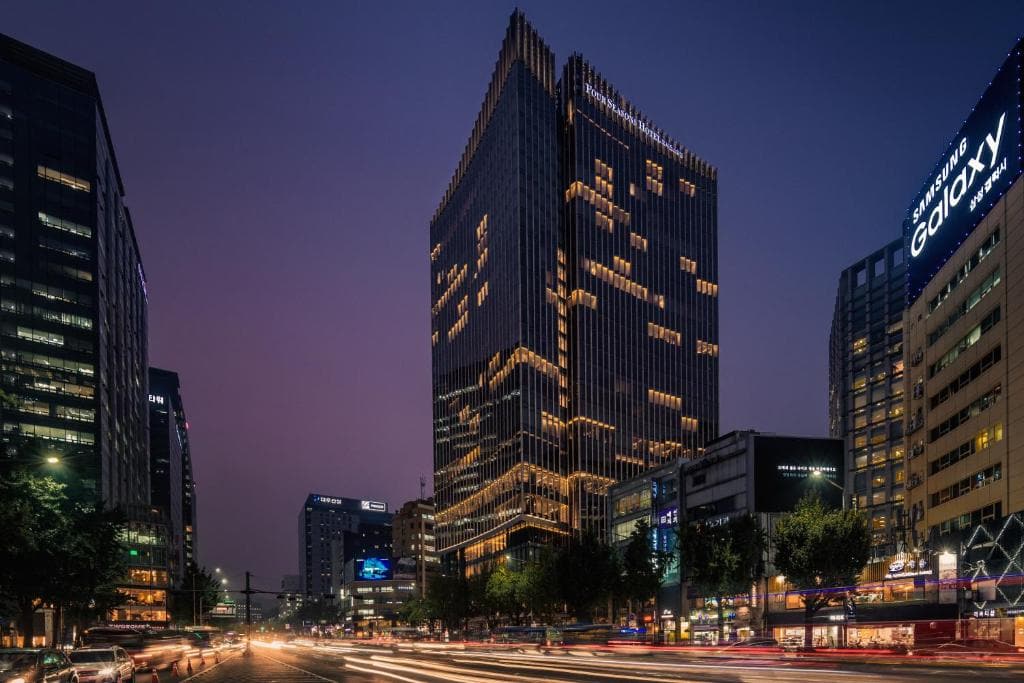 A modern cityscape at dusk shows tall office buildings with illuminated windows, a prominent glass tower in the center, and light trails from cars on a busy street below.
