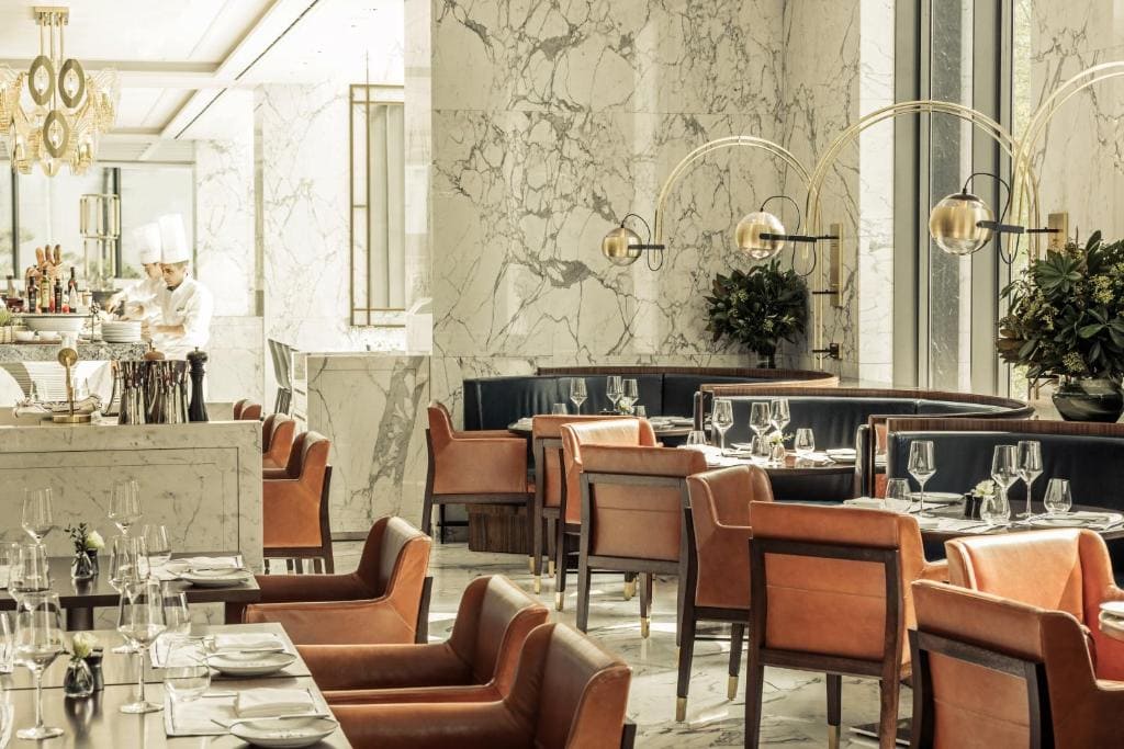 Elegant restaurant interior with marble walls, leather chairs, set tables with glassware, decorative lighting, and large windows. A chef in a white uniform works behind the counter in the background.