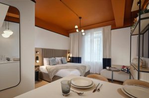 A modern studio apartment with an orange ceiling, neatly made bed, pendant lights, a dining table set for two, and a window with white and blue curtains letting in natural light.