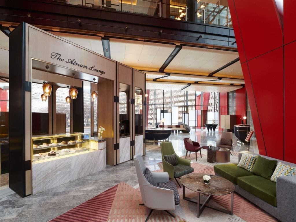A modern hotel lobby with marble floors, stylish seating areas, a green sofa, gray armchairs, and a counter labeled The Atrium Lounge. Large windows let in natural light and red structural beams decorate the space.