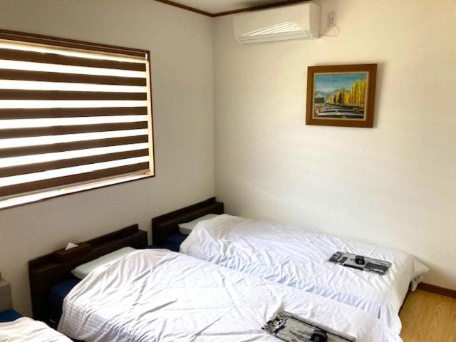 A simple room with two single beds, each with white bedding and a folded towel. A window with striped blinds, a wall-mounted air conditioner, and a framed landscape painting are visible.