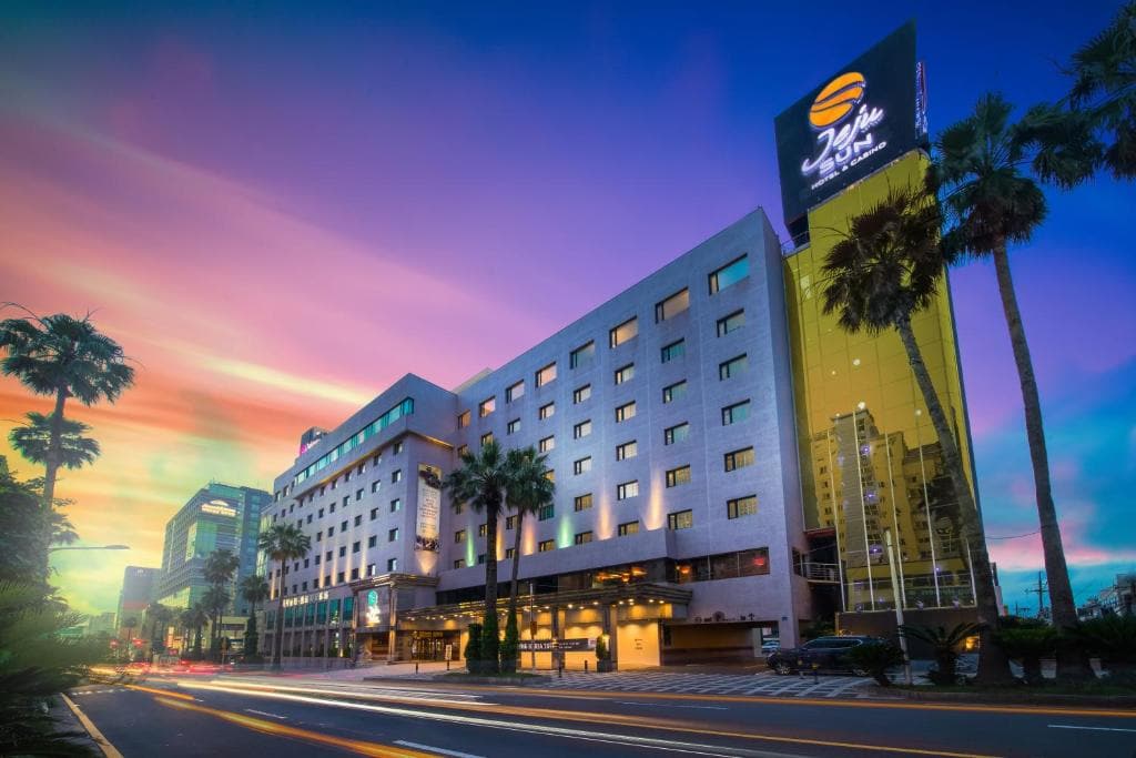 A large, modern hotel with palm trees in front, brightly lit under a colorful sunset sky. Light trails from passing cars are visible on the street, and a tall sign with a hotel logo is on the building’s corner.