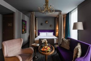 Elegant hotel room with a large bed, gold sunburst wall decor, purple velvet sofa, armchair, round table with fruit, chandelier, floor lamp, and a colorful artwork on the wall. Light streams through tall windows.