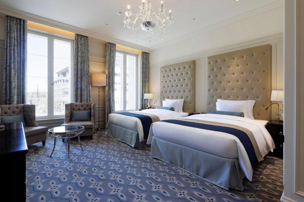 Elegant hotel room with two large beds, tufted headboards, blue and white bedding, chandelier, two armchairs by a round table, large windows with curtains, and patterned carpet.
