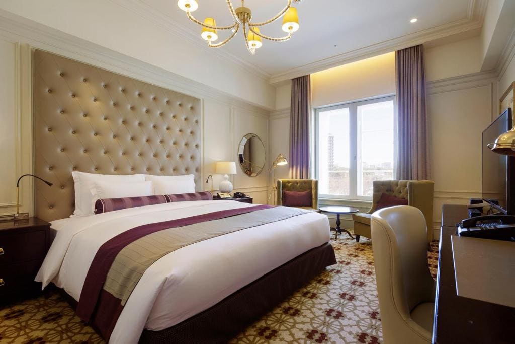 Elegant hotel room with a large bed, tufted headboard, purple and beige bedding, armchairs by a window, a desk, and a chandelier. The room is well-lit and decorated in neutral tones with gold accents.