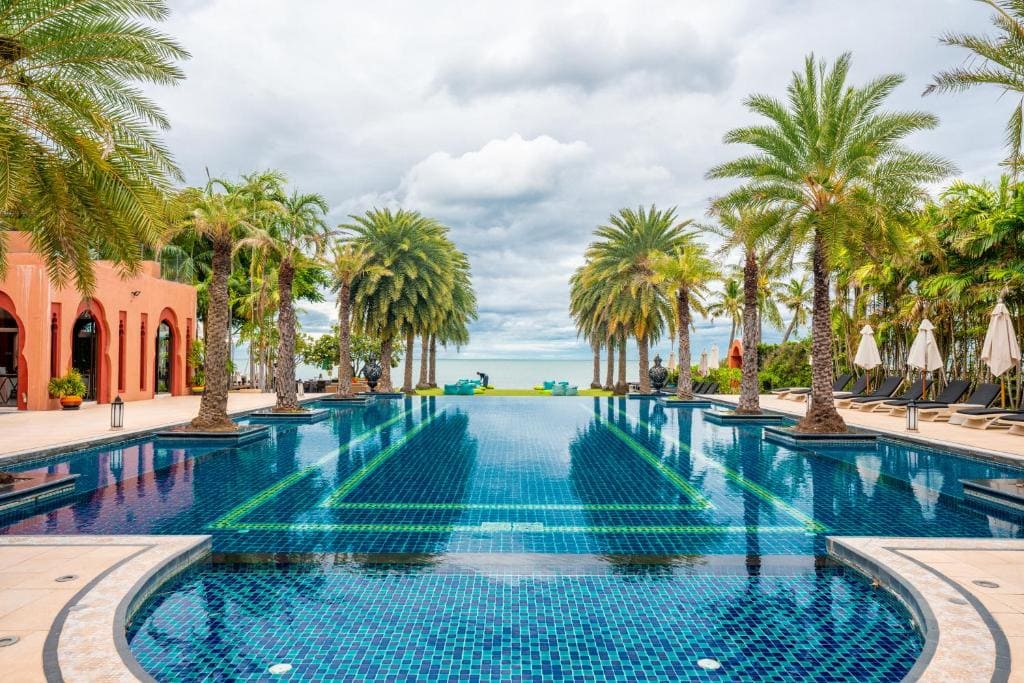 A luxurious outdoor pool surrounded by palm trees, with lounge chairs and umbrellas on either side, leads to a view of the ocean under a cloudy sky. Terracotta buildings flank the pool on the left.