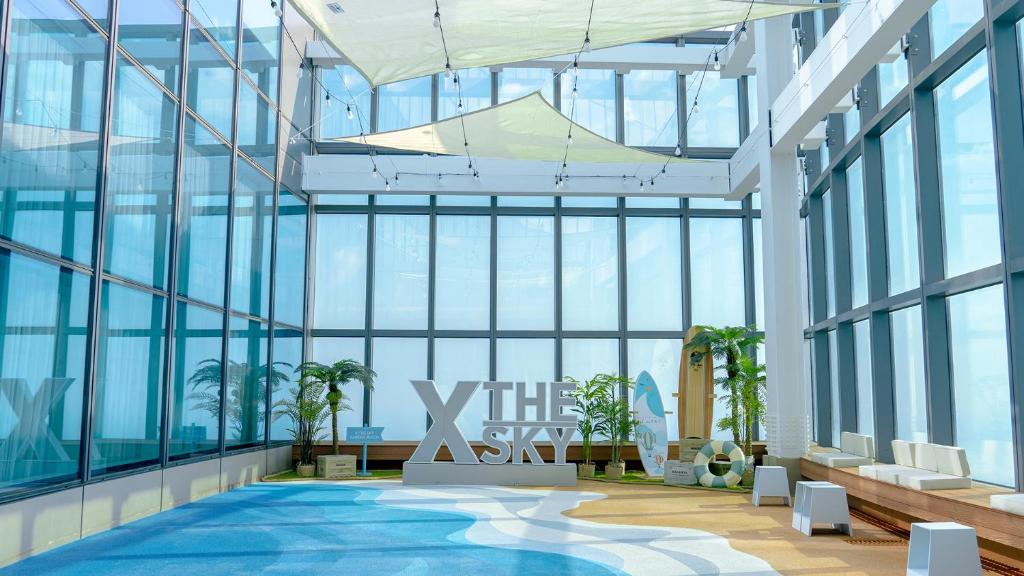 Bright indoor space with large glass windows, blue and tan flooring resembling waves, plants, white benches, and a large sign reading THE X SKY. Sunshades hang from the ceiling, creating a modern, airy atmosphere.