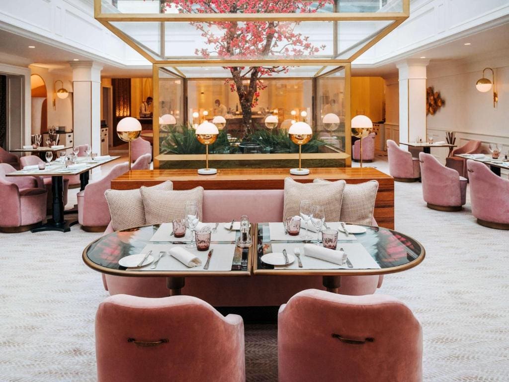 A stylish restaurant interior with plush pink chairs, set tables, and a large glass garden enclosure featuring a blooming pink tree at the center. Soft lighting and elegant decor create a modern, inviting atmosphere.