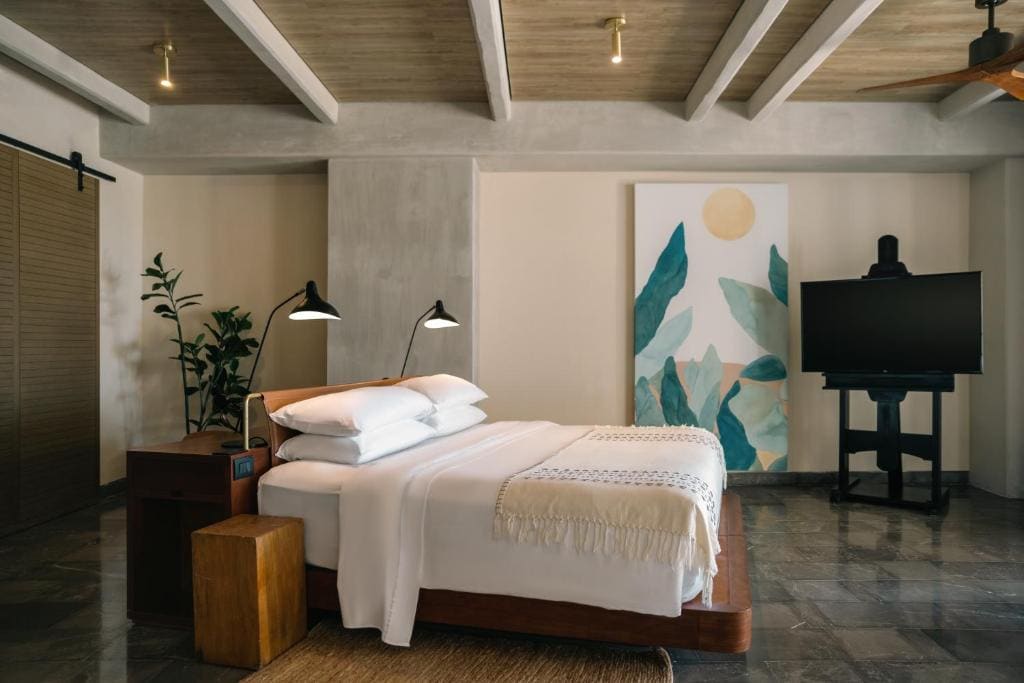 A modern bedroom with a neatly made bed, white linens, a bedside table with lamps, a potted plant, a TV on a stand, and a large abstract leaf artwork on the wall. The room has a minimalist, stylish design.