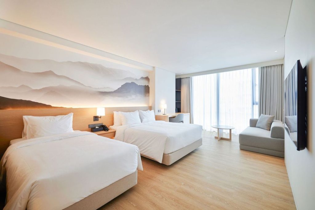 A modern hotel room with two large beds, a wall mural of mountains, a sofa, a round table, floor-to-ceiling windows with curtains, wooden flooring, and a wall-mounted TV. The space is bright and minimalistic.