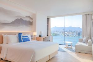 A modern, bright hotel room with a large bed, sofa, desk, and floor-to-ceiling windows offering a scenic view of a city, mountains, and a lake. Minimalist decor with light colors and a landscape mural above the bed.