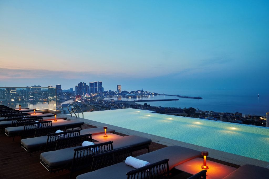 A rooftop infinity pool overlooks a coastal city at dusk, with lounge chairs and glowing lanterns lining the poolside. City lights and high-rise buildings appear in the distance near the calm sea.