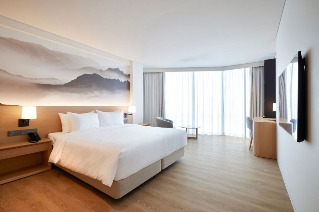 A modern hotel room with a large bed, soft white bedding, wooden floor, a mountain mural on the wall, a desk, chair, and floor-to-ceiling windows with sheer curtains letting in natural light.