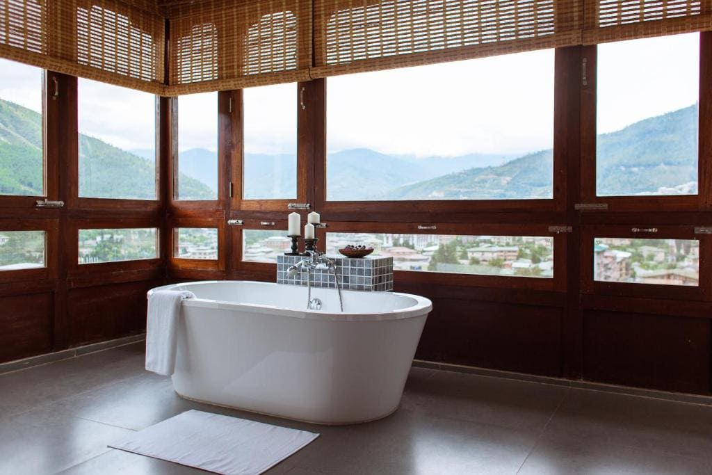 A white freestanding bathtub with a towel and mat is placed in front of large windows with wooden frames, offering a scenic mountain and valley view. Candles and bath items rest on a tray beside the tub.