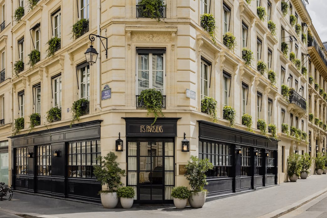 A corner view of a classic Parisian building with cream-colored walls, black window frames, and many windows adorned with green plants. The ground floor houses a restaurant named “Les Parisiens.”.