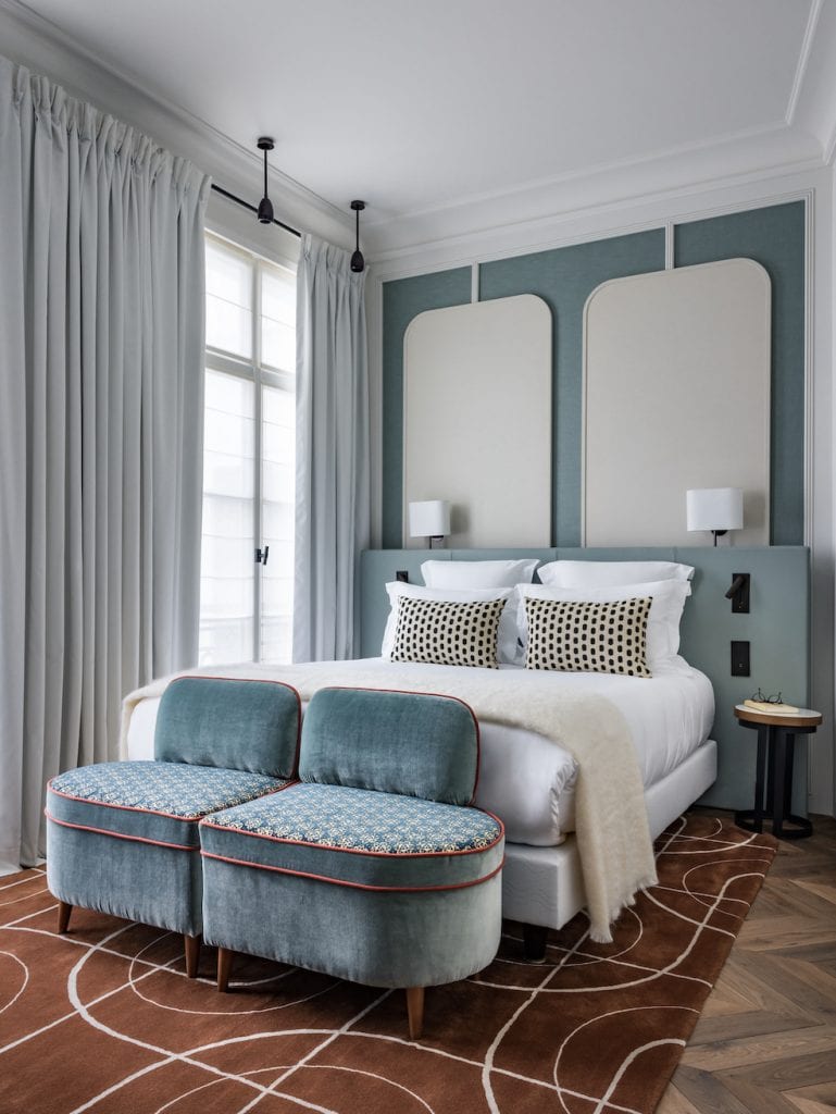 A modern bedroom with a double bed, two blue upholstered benches at the foot, patterned cushions, tall curtains, wall-mounted lamps, and a geometric brown rug on a wooden floor.