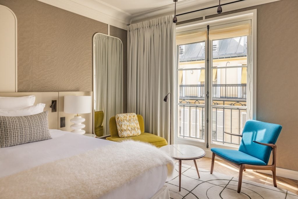 A bright hotel room with a large bed, a white lamp, an oval mirror, a yellow armchair, a blue chair, a small round table, and a window with sheer curtains overlooking balconies.