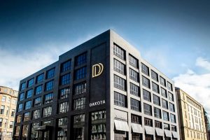 A modern, dark-gray five-story building with large windows and a gold D logo on the corner, labeled Dakota. The sky is blue with clouds, and neighboring buildings are visible.