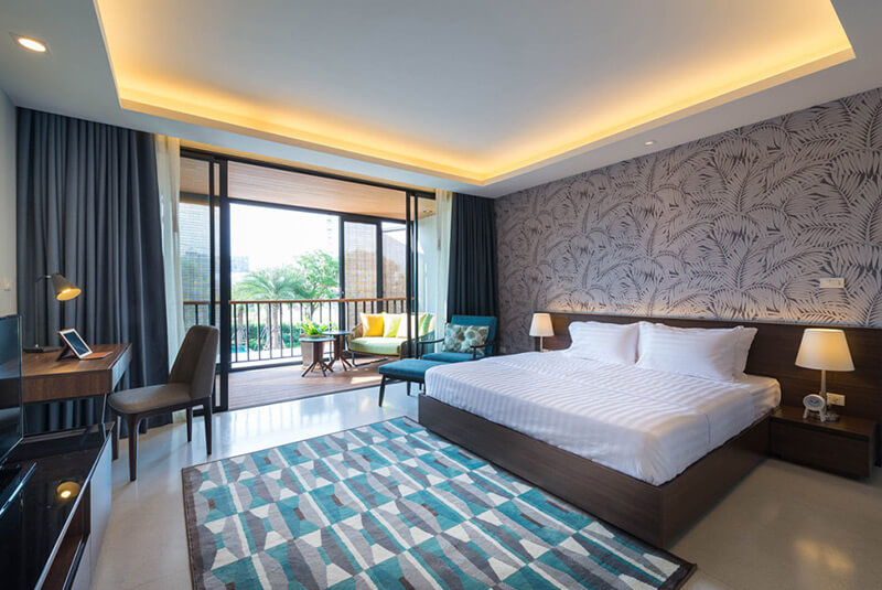 Modern hotel room with a large bed, patterned wallpaper, desk, TV, and balcony featuring outdoor seating. Room is bright with natural light and decorated in neutral and blue tones.