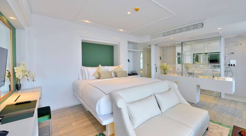 A modern hotel room with a large bed, green accent wall, white sofa, desk, TV, and an open-plan bathroom with glass walls, double sinks, and bright lighting. The décor is minimalist with light colors.