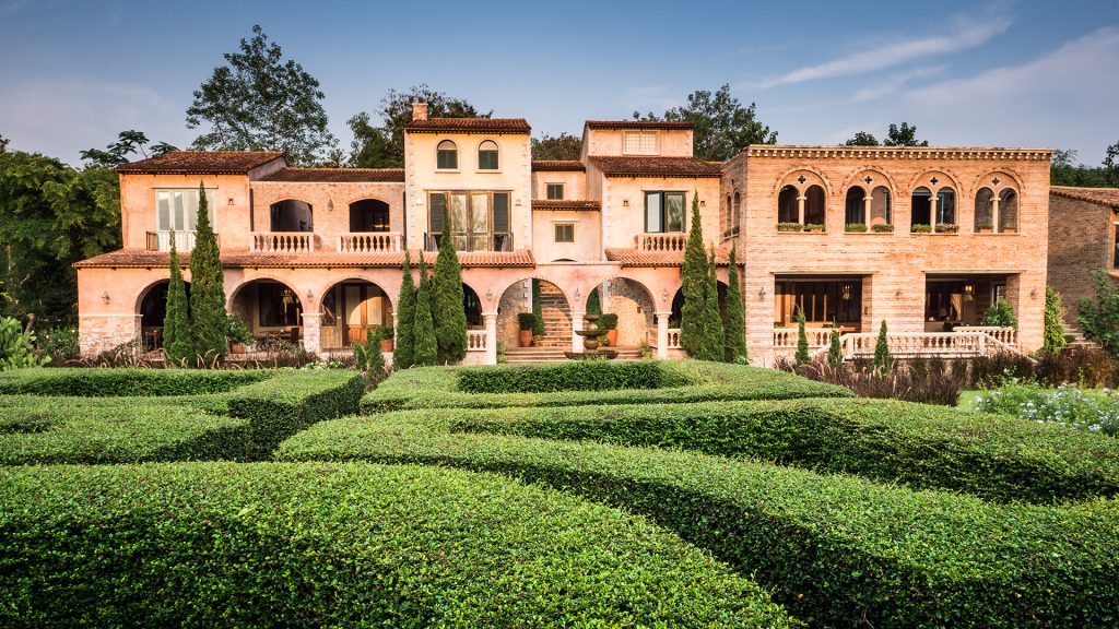 A large, Mediterranean-style villa with arched windows, terracotta roofs, and balconies, surrounded by neatly trimmed hedges and lush greenery under a clear blue sky.