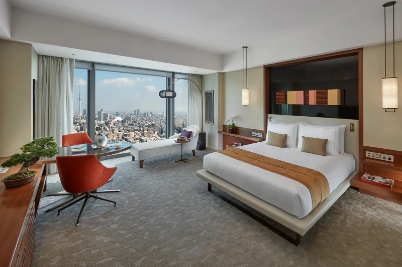 Modern hotel room with large bed, orange chairs, a chaise lounge by a floor-to-ceiling window, and a stunning city skyline view. Warm tones, soft lighting, and minimalist decor create a comfortable, inviting atmosphere.