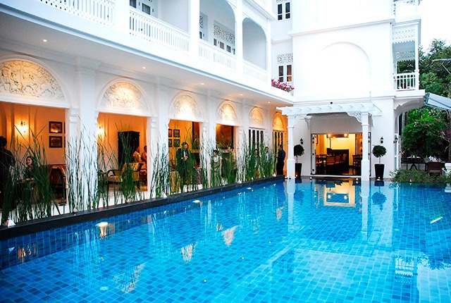 A luxurious hotel courtyard with a blue-tiled swimming pool, surrounded by white colonial-style architecture, arches, and outdoor lighting. Green plants line the edge of the pool and balconies overlook the area.