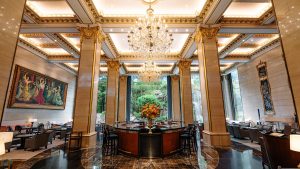 A grand hotel lobby with tall marble columns, ornate gold detailing, chandeliers, and large windows overlooking lush greenery. Elegant seating areas, a central round table with a floral arrangement, and framed art decorate the space.