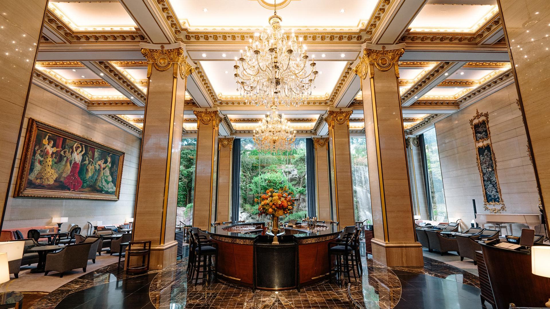 A grand hotel lobby with tall marble columns, ornate gold detailing, chandeliers, and large windows overlooking lush greenery. Elegant seating areas, a central round table with a floral arrangement, and framed art decorate the space.