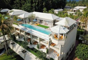 Aerial view of a luxurious, multi-story villa with a long infinity pool, sun loungers, manicured lawns, and open-air lounge areas surrounded by tropical trees.