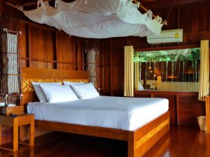 A cozy bedroom with wooden walls and flooring, featuring a double bed with white linens, a canopy net overhead, bedside tables, curtains, and a large window overlooking greenery. An air conditioner is mounted above the window.