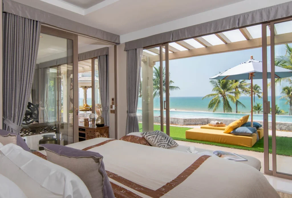 A bright, modern bedroom with a large bed opens onto a patio with a sunbed, umbrella, green lawn, palm trees, and a scenic ocean view under a clear sky.