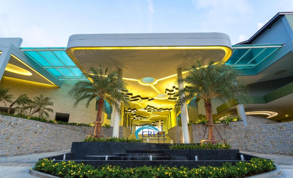 Modern building entrance with yellow-lit ceiling, abstract light fixtures, palm trees, stone walls, and a water fountain surrounded by greenery and yellow flowers. The CREST sign is visible near the entrance.