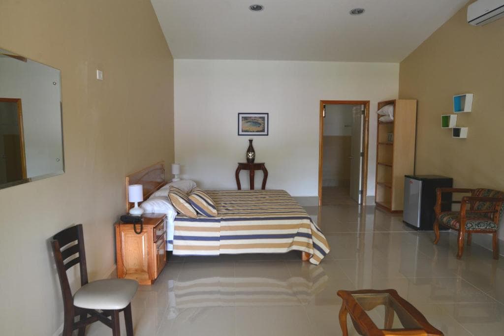 A spacious, tidy bedroom with a striped bedspread, wooden furniture, a wall-mounted TV, and an open shelf. The room features tiled floors, light walls, and a small table with two chairs.