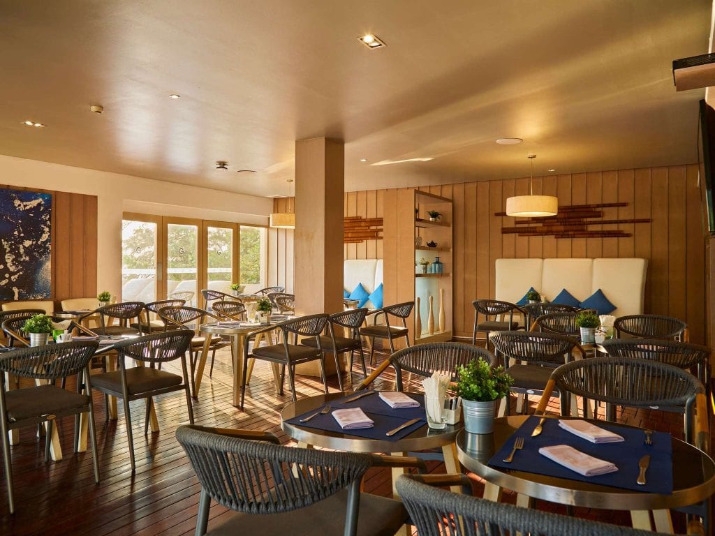 Modern restaurant interior with wooden flooring, round tables set with blue placemats, and woven chairs. Large windows let in natural light, and decorative plants and wall art add to the cozy, contemporary ambiance.