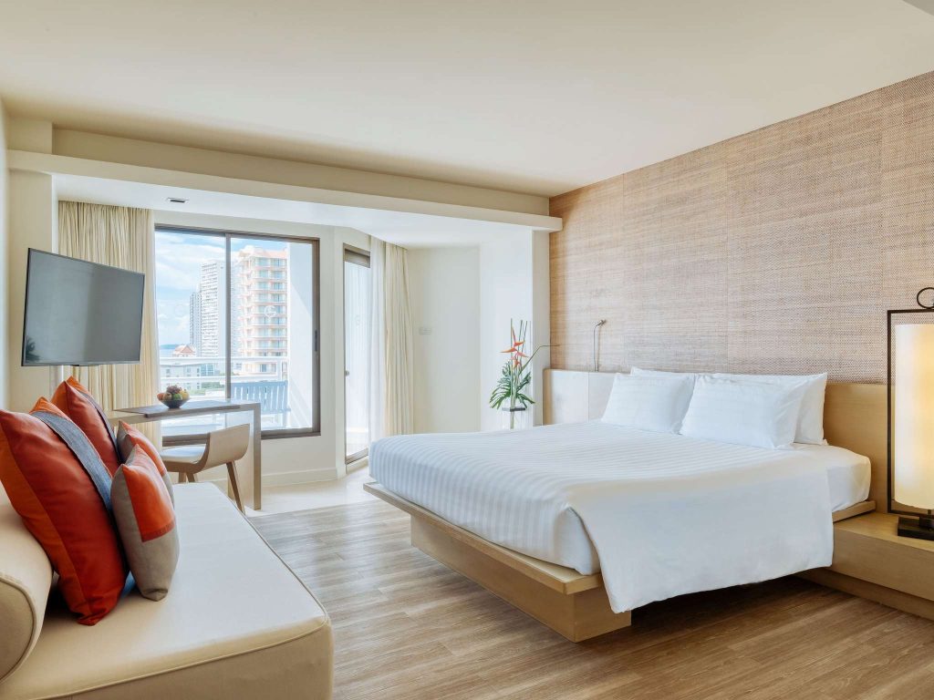 A modern hotel room with a large bed, white linens, a beige sofa with orange and gray pillows, a flat-screen TV, wood flooring, a large window, and a city view with tall buildings.