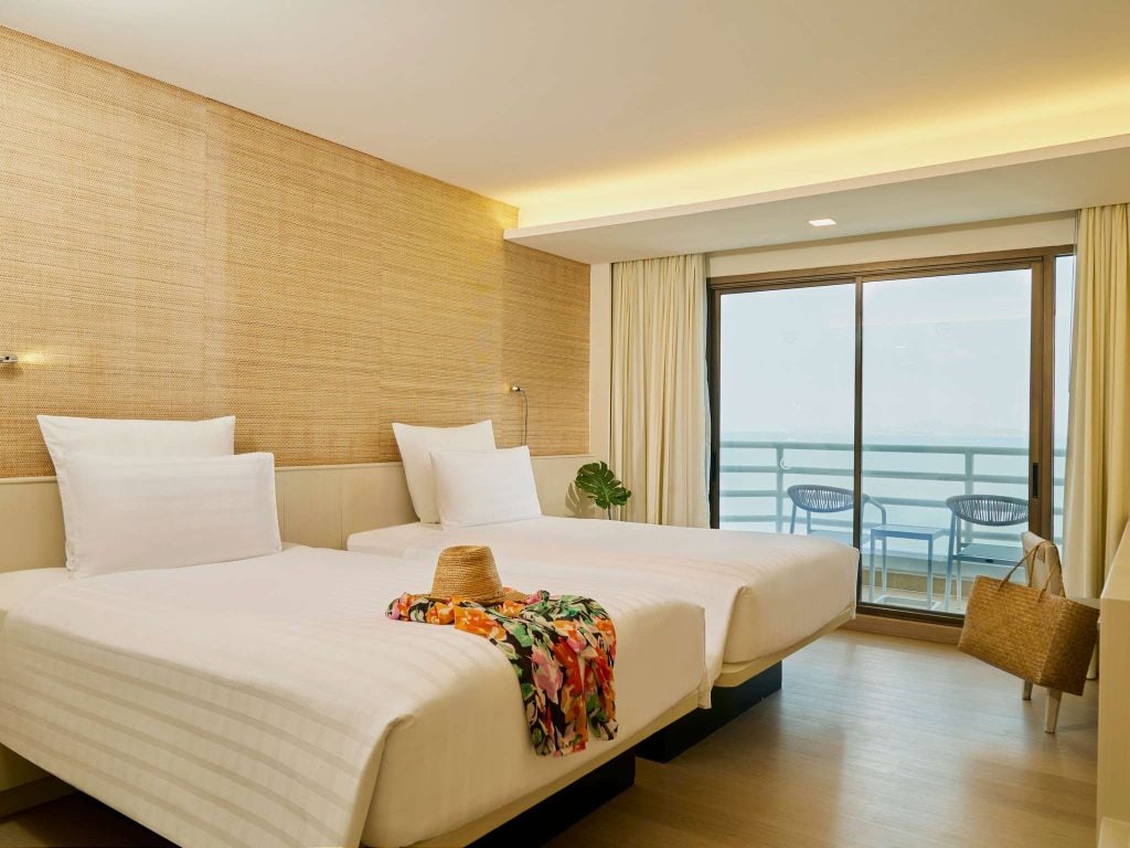 A bright hotel room with two white twin beds, a floral dress and straw hat on one, a sliding glass door leading to a balcony with chairs, and a woven bag hanging on the wall.