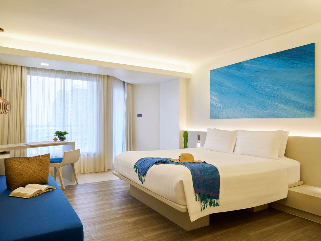 A modern hotel room with a large bed, white bedding, blue throw, and hat on the bed. Theres a blue sofa, an open book, a desk with a chair by a large window, and a large abstract blue painting on the wall.