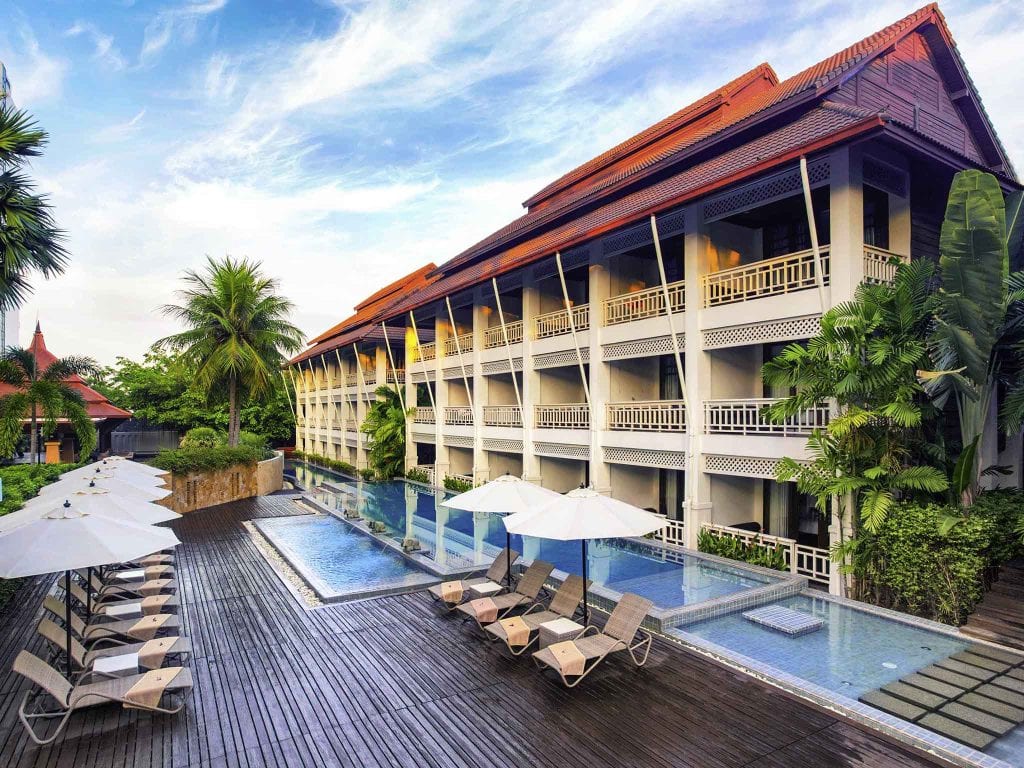 A tropical resort with a large pool, lounge chairs and umbrellas on a wooden deck, surrounded by palm trees and lush greenery, with a three-story building featuring balconies in the background under a blue sky.