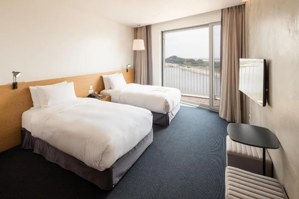 Modern hotel room with two single beds, a large window overlooking a balcony with a scenic view, wall-mounted TV, bedside table, and minimalist decor in neutral tones.