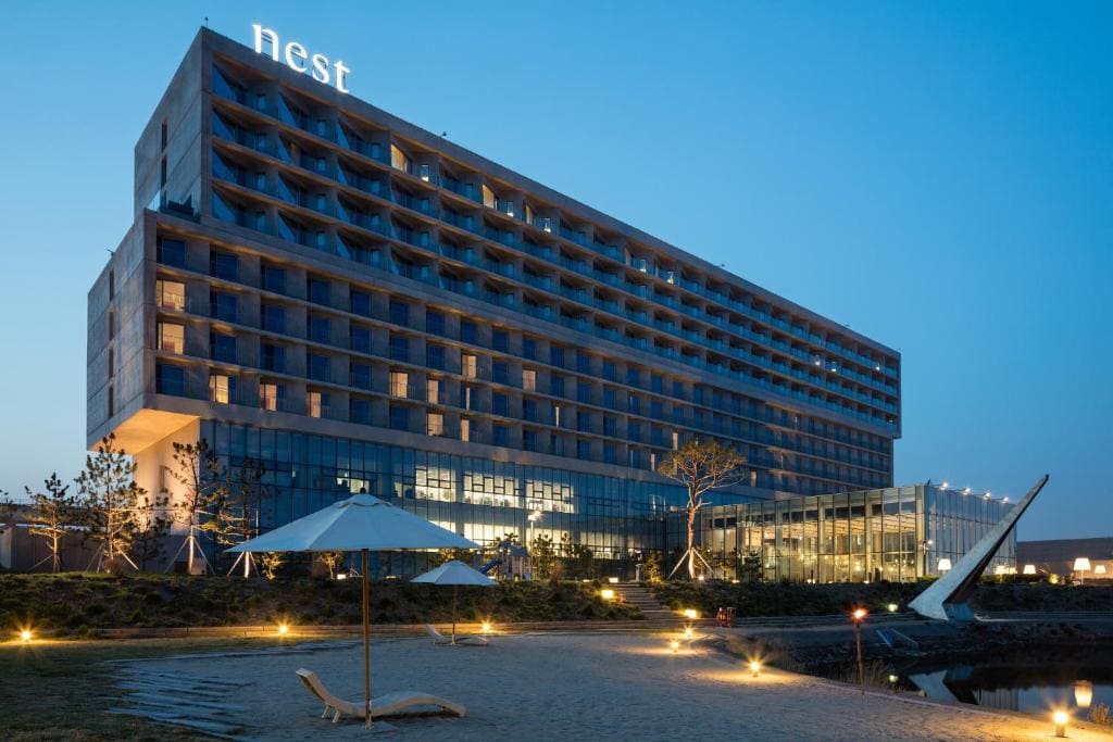 Modern multi-story hotel with the word nest lit on the roof, features glass and concrete design, set against a clear evening sky. A sandy area with sun loungers and umbrellas is in the foreground.