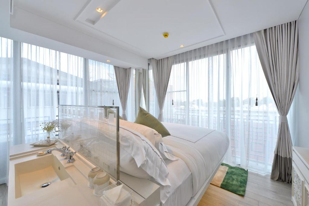 A modern, bright hotel room with large floor-to-ceiling windows, sheer curtains, a neatly made bed with white linens, and a sink with toiletries next to the bed. Natural light fills the space.
