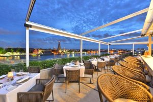 Rooftop restaurant with woven chairs and white tablecloths overlooking a river at sunset, cityscape and illuminated buildings visible in the background under a blue, cloud-filled sky.