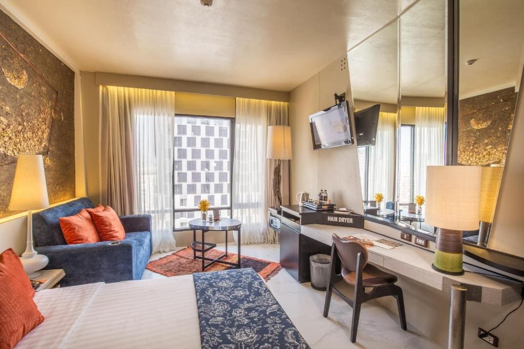 A modern hotel room with a bed, blue sofa, and orange cushions, a small round table by a window, a desk with a chair, a wall-mounted TV, and warm lighting highlighting gold-accented decor.