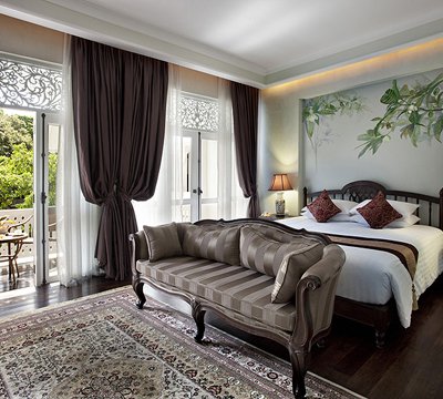 Elegant bedroom with a striped sofa, a large bed with pillows, dark curtains, a patterned rug, and a balcony with greenery visible through glass doors. The walls feature a floral mural above the bed.