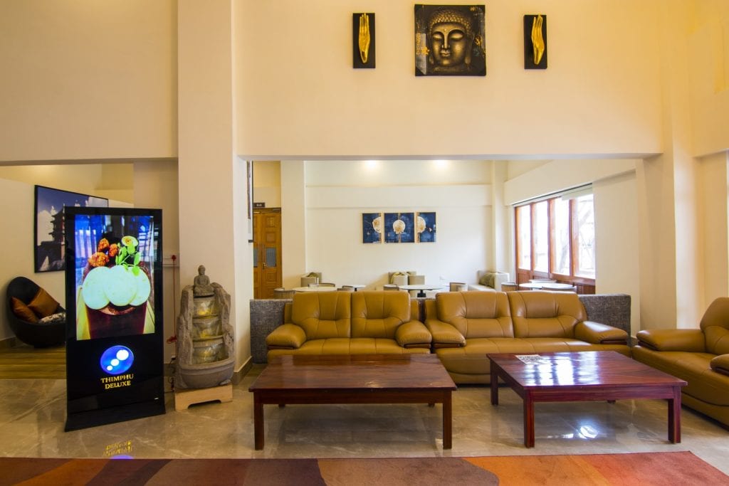 A modern lounge area with tan leather sofas, wooden coffee tables, a floor statue, digital display screen, and wall art including a Buddha painting and decorative panels. Large windows bring in natural light.