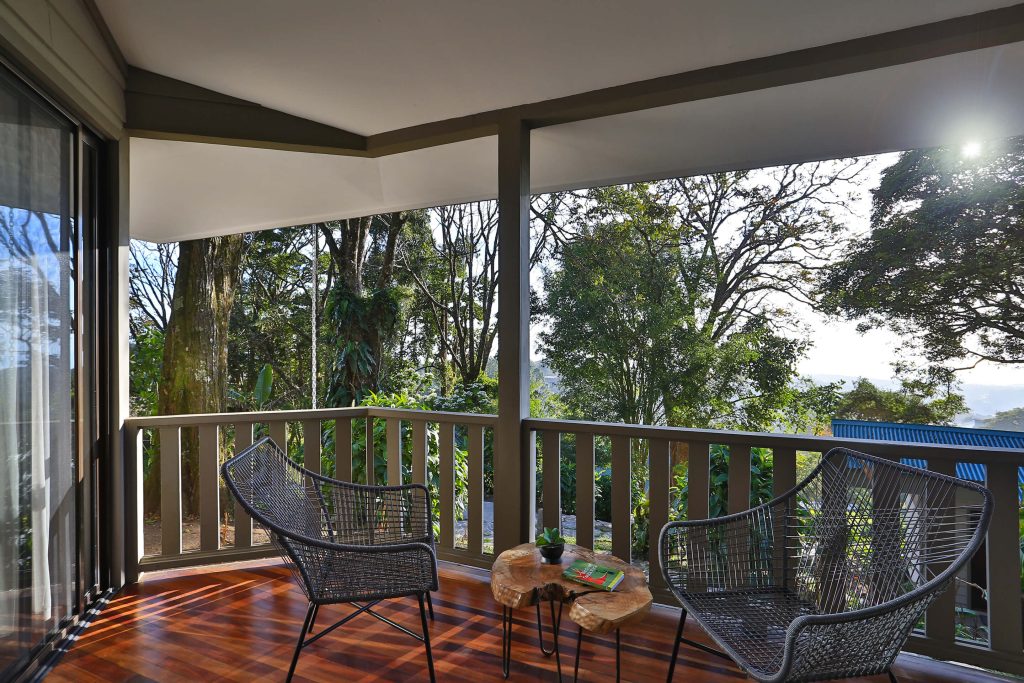 A cozy balcony with two modern chairs and a small wooden table overlooks a lush, green forest. Sunlight filters through the trees, creating a peaceful and inviting outdoor space.